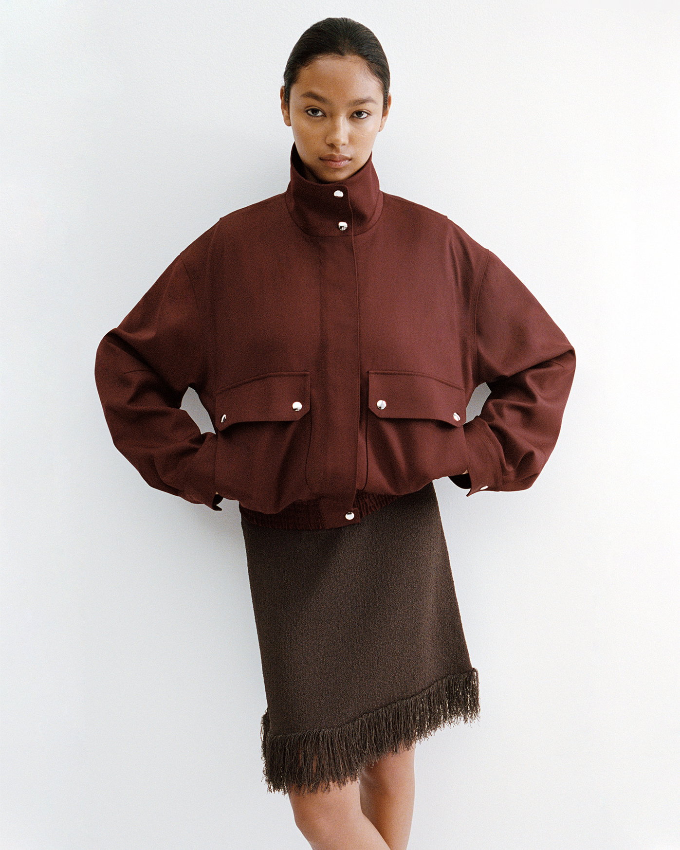 Cropped image of model standing against a white wall wearing Saski Dress in Cotton in Textured Fringe in Earth, with the Aviva Jacket in Wool Gabardine in Oxblood layered over