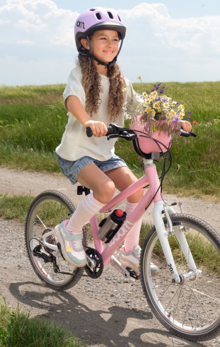 A girl rides a powder pink woom GO 4 down a dirt road, complete with a POP Basket full of wildflowers