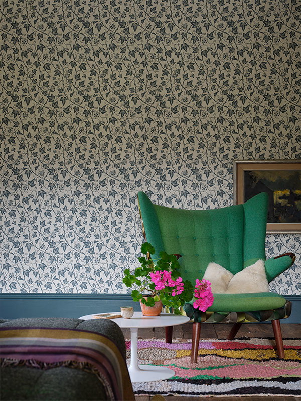 A green, upholstered armchair in front of an Ivy wallpaper in blue and white. The pattern is small and repeats often, creating a busy feel that draws your attention. The space feels modern style and as though each element has been carefully curated.