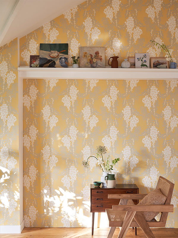 A bright sitting room decorated with yellow Wisteria wallpaper and a wooden armchair. The space feels warm and inviting, as though you could sit down and enjoy sunlight on your face.
