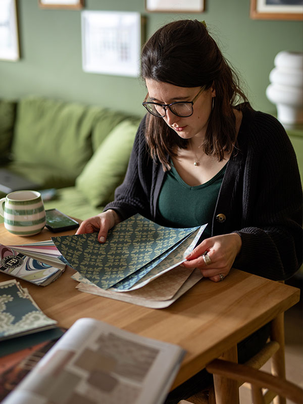 A brunette woman holding a selection of A4 wallpaper samples. She’s sat at a wooden table, which also has more samples, sample pots and a colour card on. In the background is an olive-green wall, dresser and sofa all the same shade.
