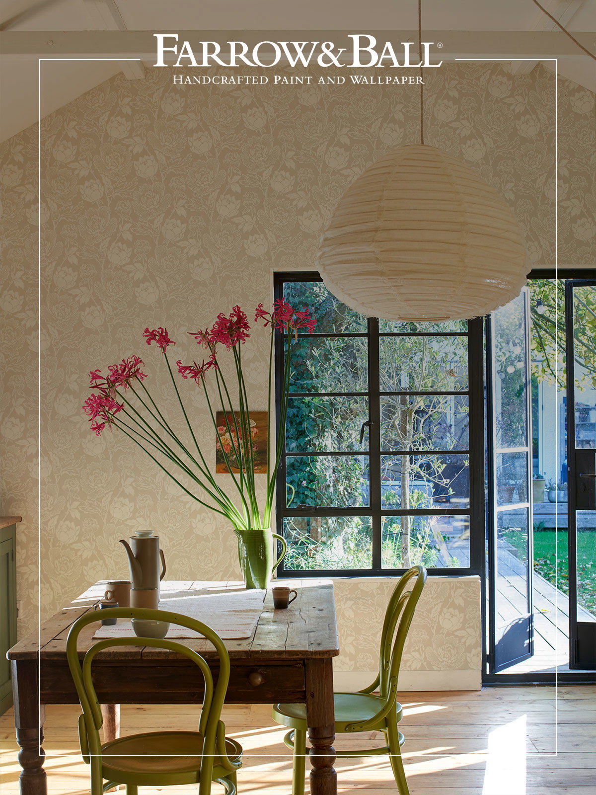 A calming, open kitchen with Peony wallpaper in a soft, neutral colourway. There is a wooden table in front of the wall and an open glass door, all creating a relaxed, inviting feel.