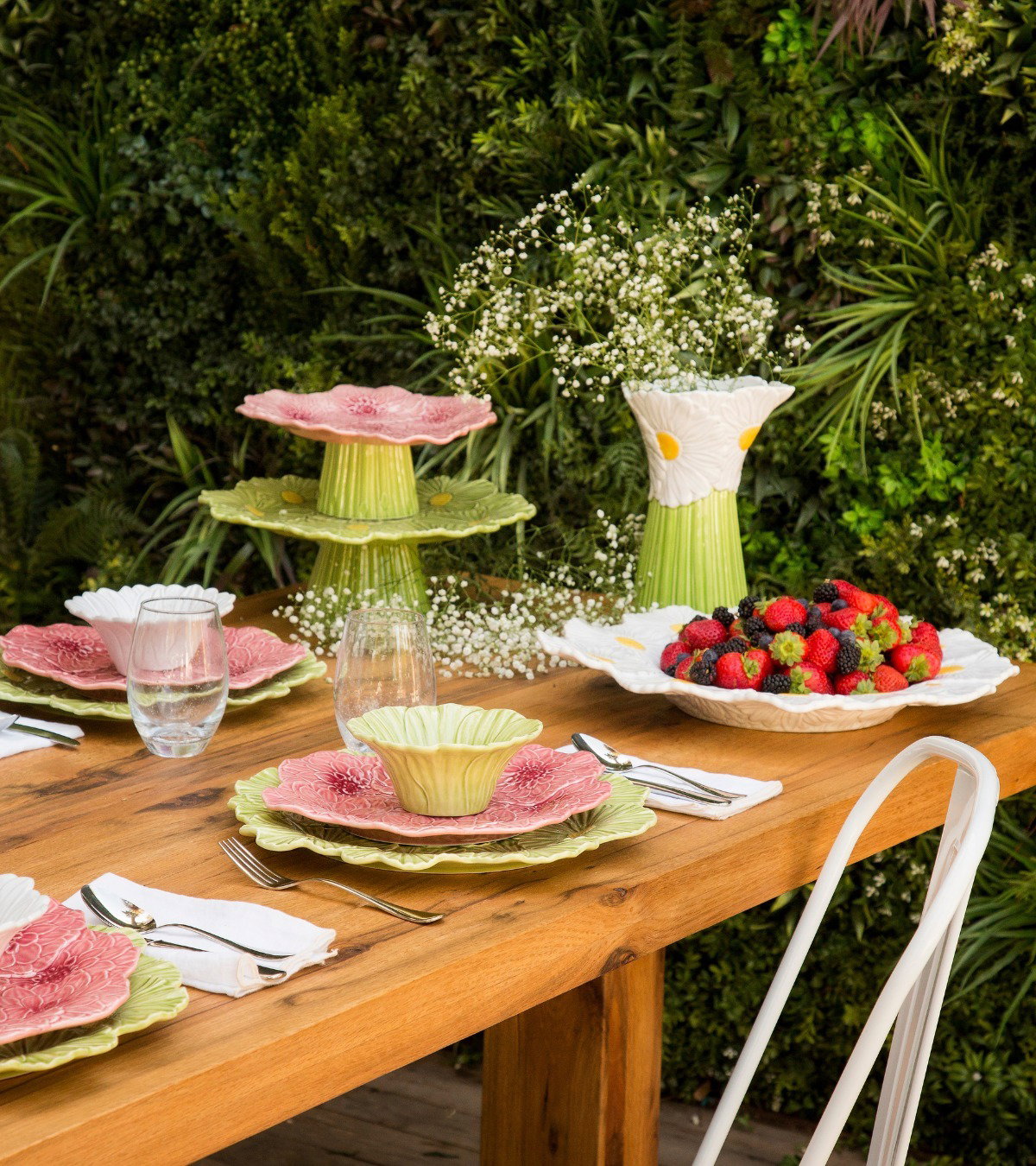 Outdoor wooden dining table set with floral ceramic plates, cake stands and vase, bowl of mixed berries against a green living wall