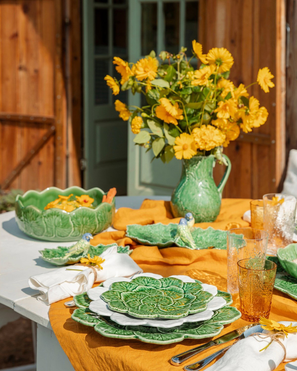 Sunlit outdoor table with green leaf-pattern plates and bowls, mustard cloth, yellow flowers in a green jug and amber glasses