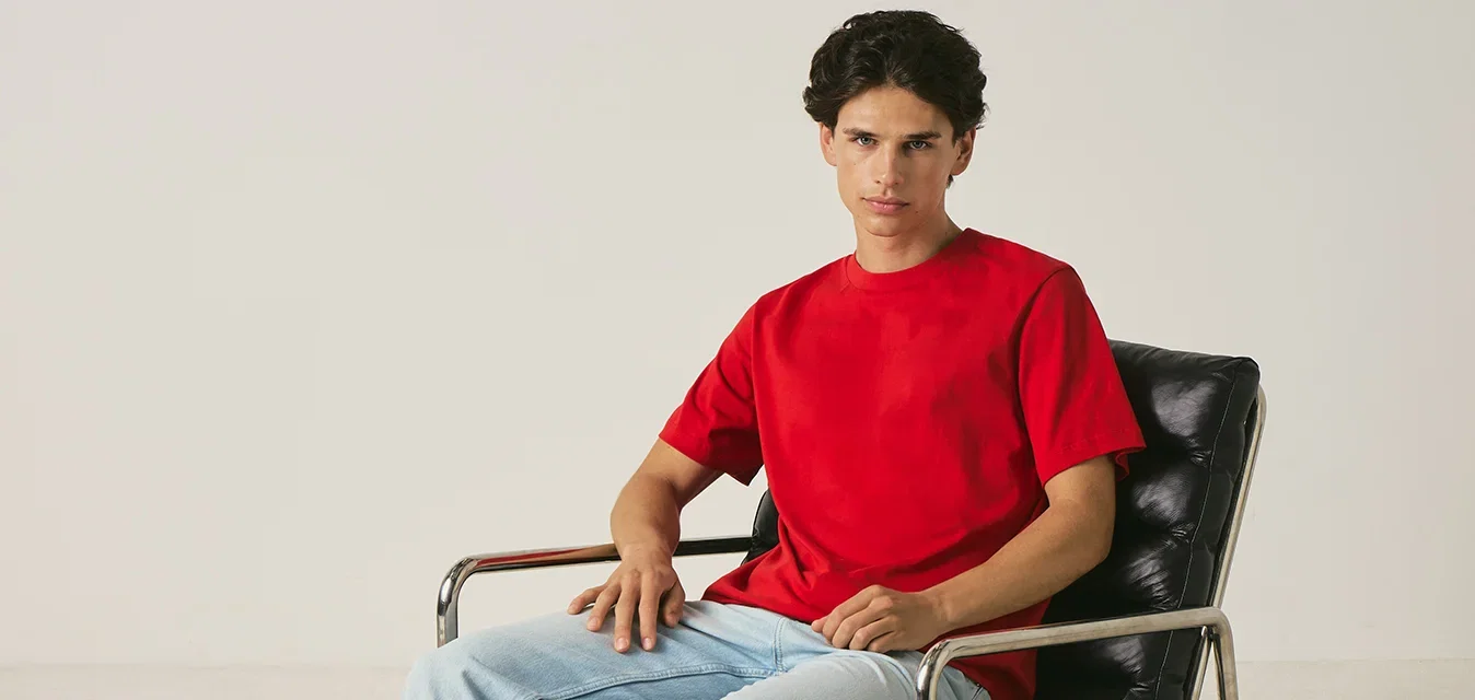 Joven con camisa roja y vaqueros claros sentado en una silla de cuero negra con brazos metálicos sobre un fondo claro y liso.