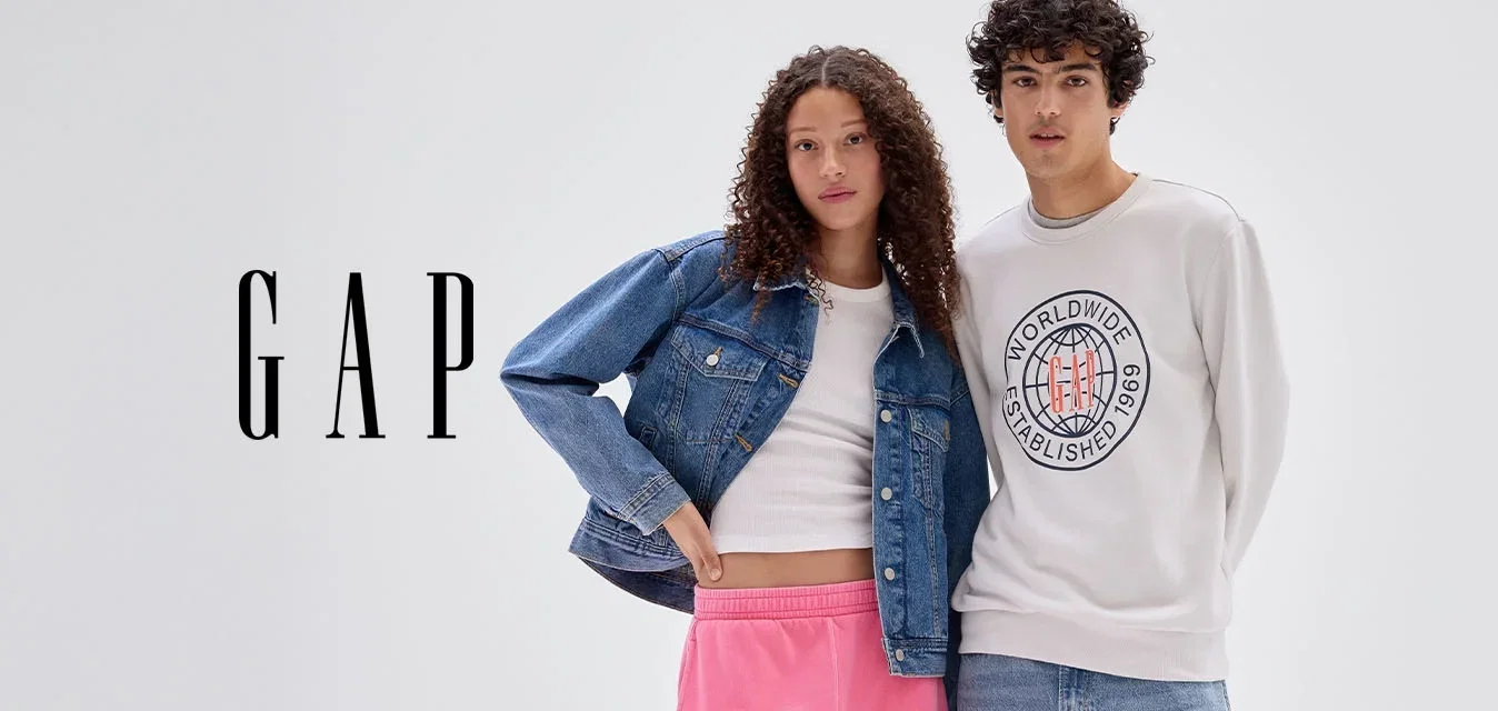Dos jóvenes de pie juntos, mujer con pantalones deportivos rosas y chaqueta vaquera, hombre con sudadera clara y vaqueros, fondo gris neutro.