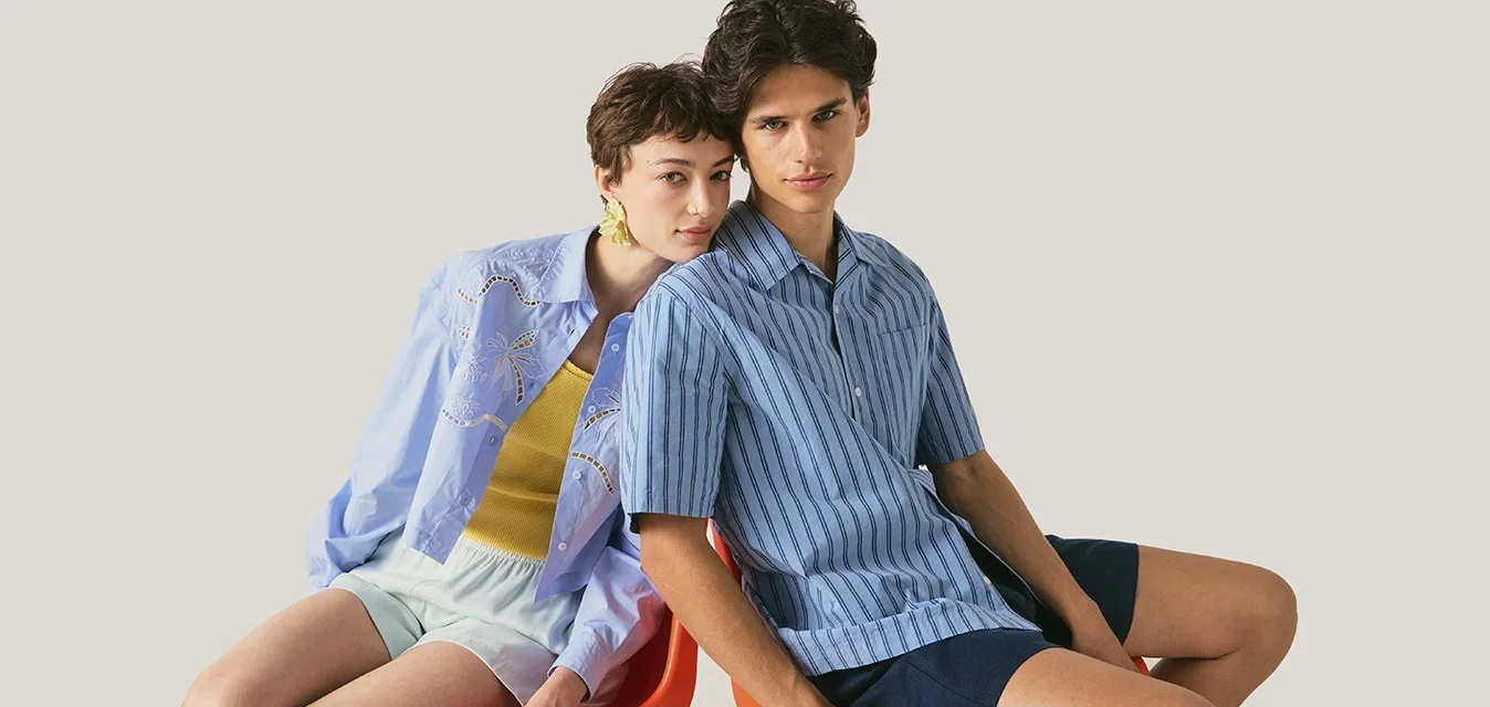 Dos personas sentadas juntas en una silla roja, una lleva una camisa azul a rayas y pantalones cortos oscuros, la otra una camisa azul claro con pantalones cortos blancos.