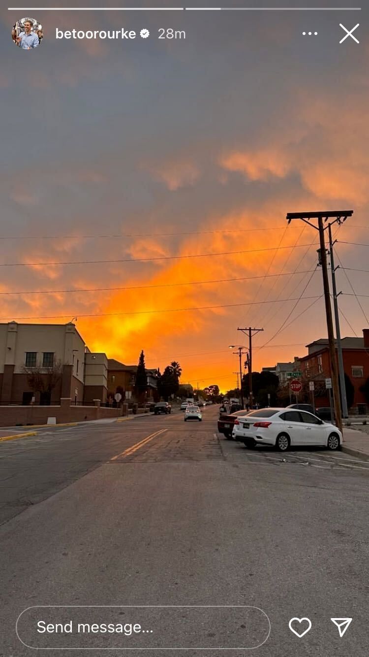 Screenshot from @betoorourke Instagram story of a sunset over a city street, with multiple cars parked and one car driving ahead, surrounded by buildings