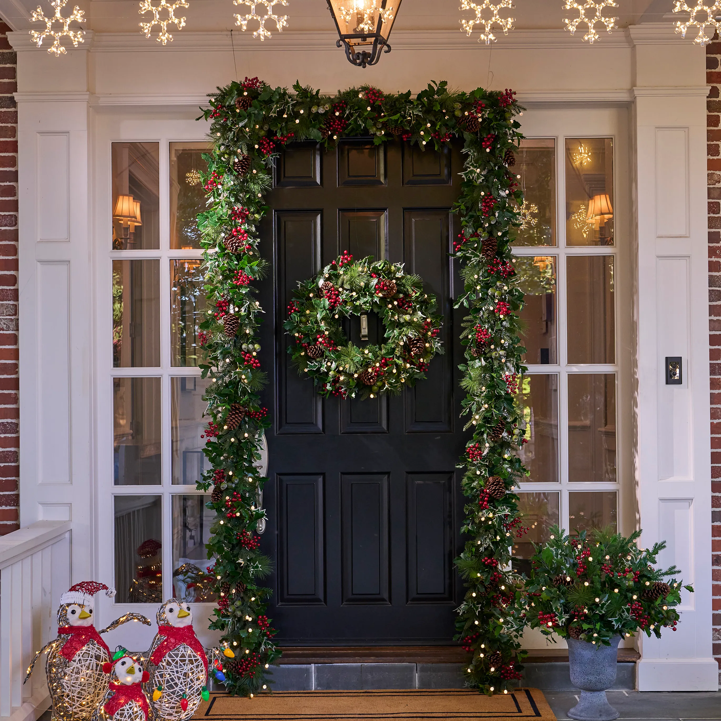 Pre-lit Christmas garland and wreath with red berries and pinecones decorating a black front door, with illuminated penguin figures by the entrance.