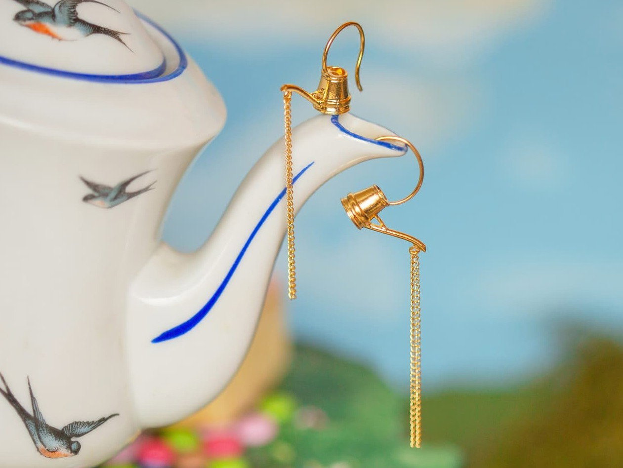Alex Monroe Gold Watering Can Pouring Chain Hook Earrings with delicate chain detail styled beside a white teapot.
