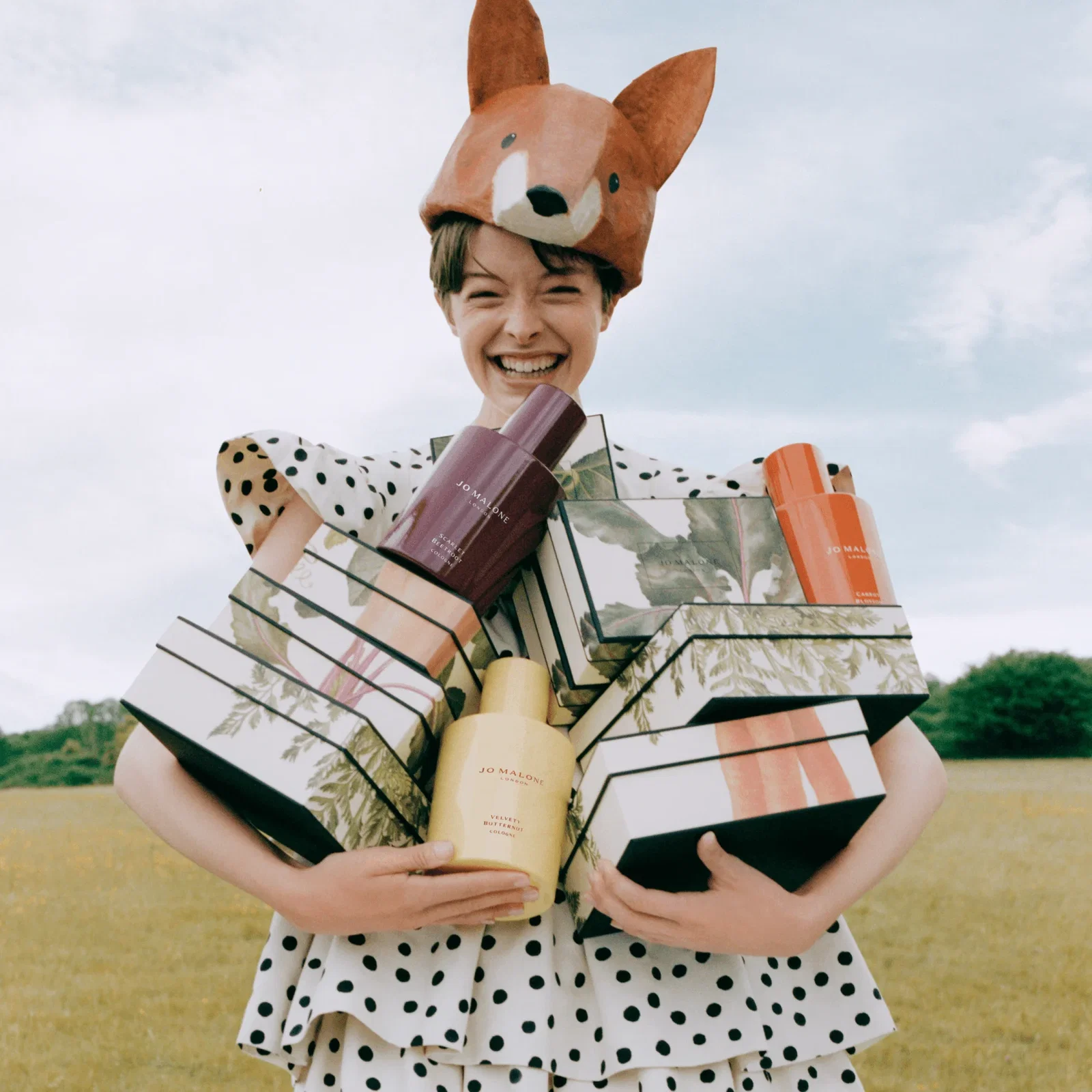 Woman standing in a field, smiling, wearing a large polka-dotted dress and a fox hat, holding Jo Malone boxes and cologne bottles from the Veggies collection.