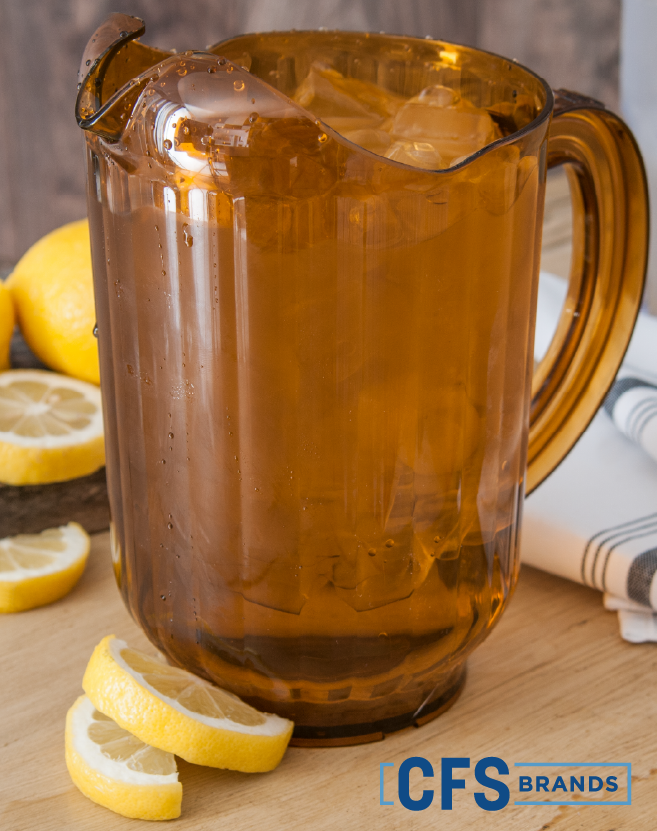 CFS Brands Carlisle amber beverage pitcher filled with ice, displayed on a wooden surface with fresh lemon slices and a folded striped napkin.