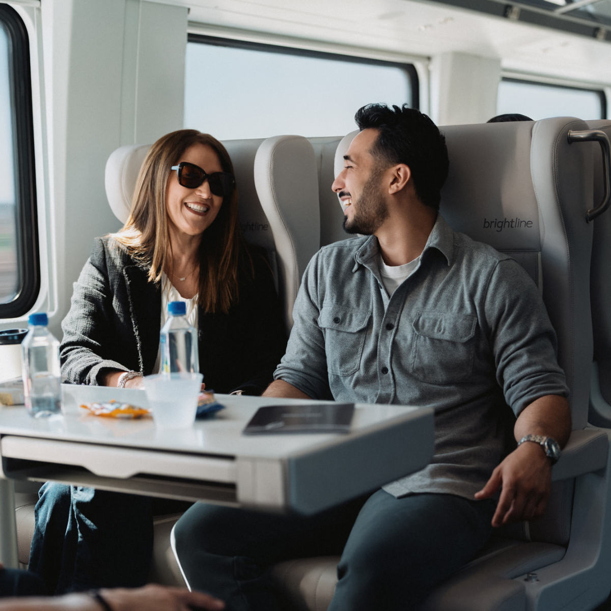 A man and woman sit together at a table on a modern train, smiling and talking. They are seated in comfortable high-backed seats with large windows showing a blurred outdoor landscape. A water bottle, cup, and snacks rest on the table between them, suggesting a relaxed journey. Soft daylight and neutral tones create a warm, casual travel atmosphere.