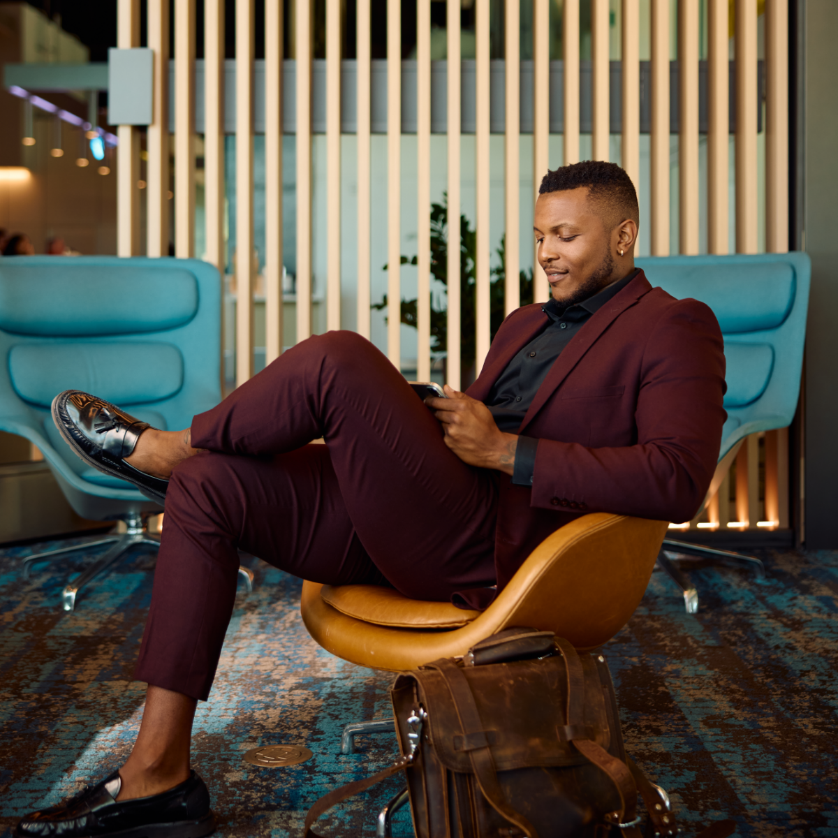 A well-dressed man in a burgundy suit sits in a modern lounge chair, focused on his smartphone. He appears to be in a contemporary office or airport lounge with stylish blue armchairs and vertical wooden slats in the background. A brown leather bag rests on the floor beside him, suggesting travel or commuting. The scene feels relaxed yet professional, with warm lighting and a clean, modern interior design.