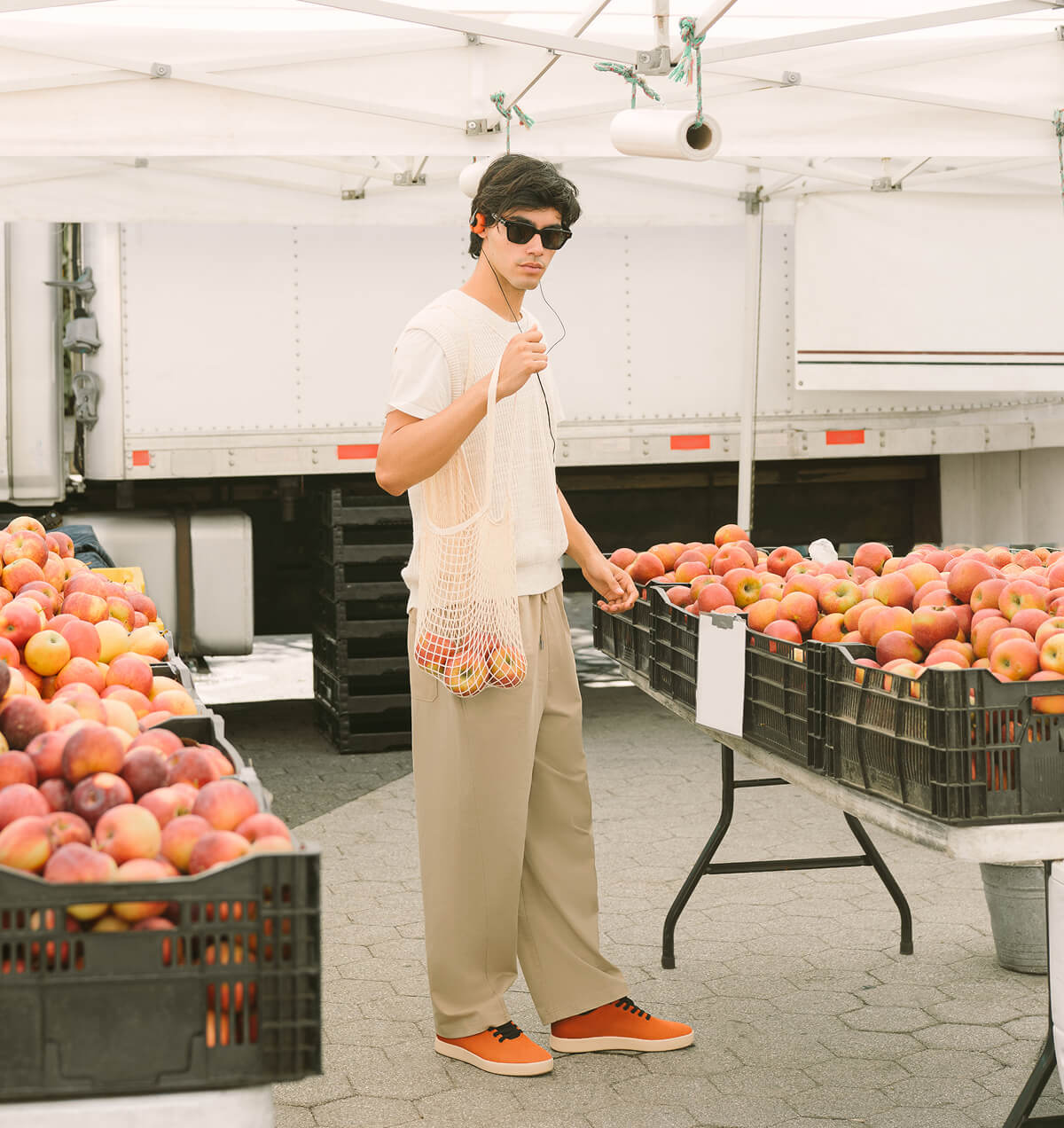 "Man in sunglasses shops for apples at outdoor market, holding a mesh bag. Crates of apples surround him under a white canopy."