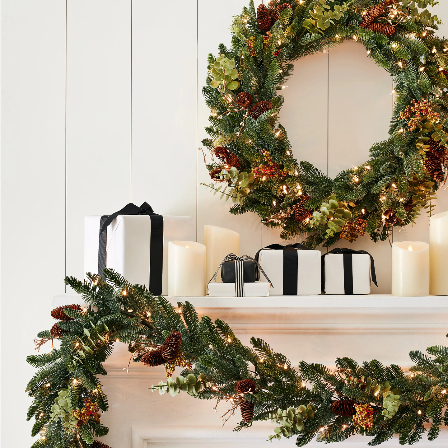 Pre-lit Christmas wreath and garland with pinecones, greenery, and warm lights displayed on a white mantel with candles and wrapped gifts.