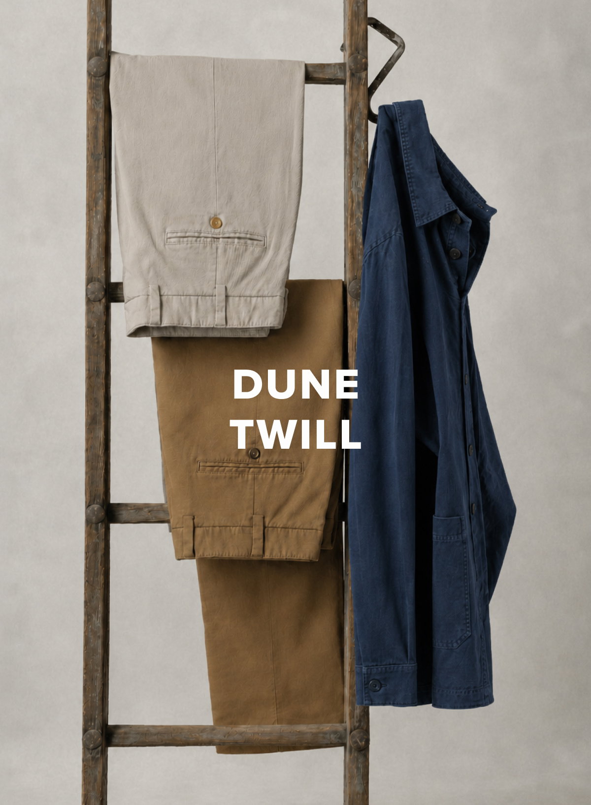 Two pairs of Bills Khakis Dune Twill pants and a Dune Twill Chore Jacket hand from a vintage ladder against a beige backdrop. Text reads Dune Twill
