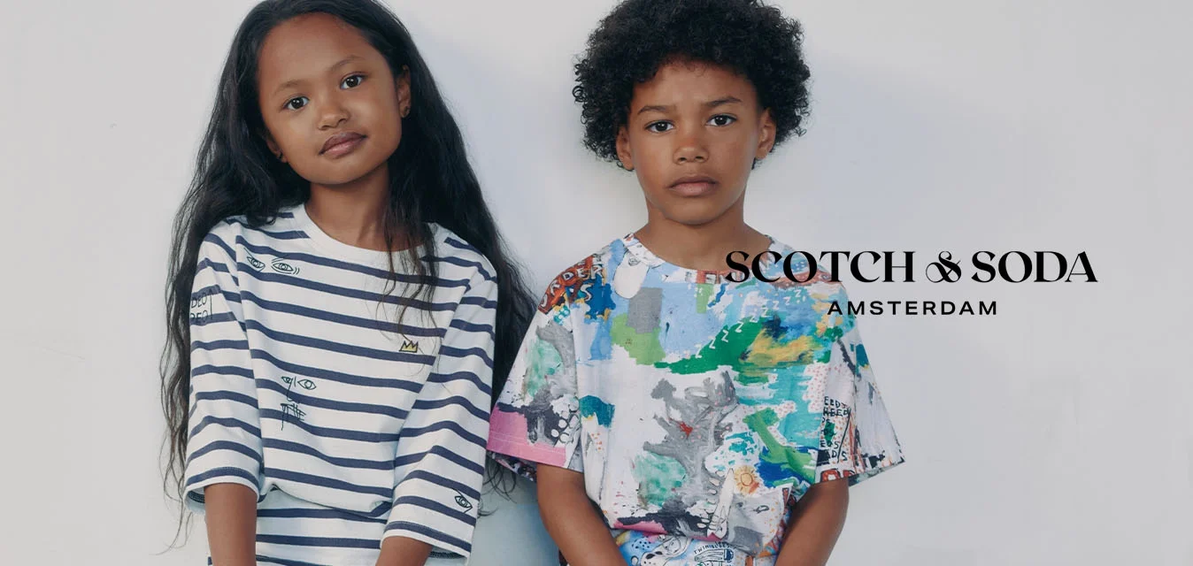 Twee kinderen zitten naast elkaar tegen een witte muur; het meisje draagt een gestreept shirt en witte sneakers, de jongen draagt een kleurrijk T-shirt en beige schoenen.