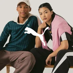 Twee modellen zitten op een houten bank. De man draagt een groen gestreept shirt en een paarse broek, terwijl de vrouw een zwarte jumpsuit met roze accenten heeft.