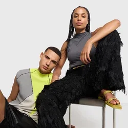 Due modelli in abbigliamento moderno posano di fronte a uno sfondo neutro; uno è seduto su una sedia con pantaloni neri e sandali gialli, l'altro è sdraiato accanto a lui.
