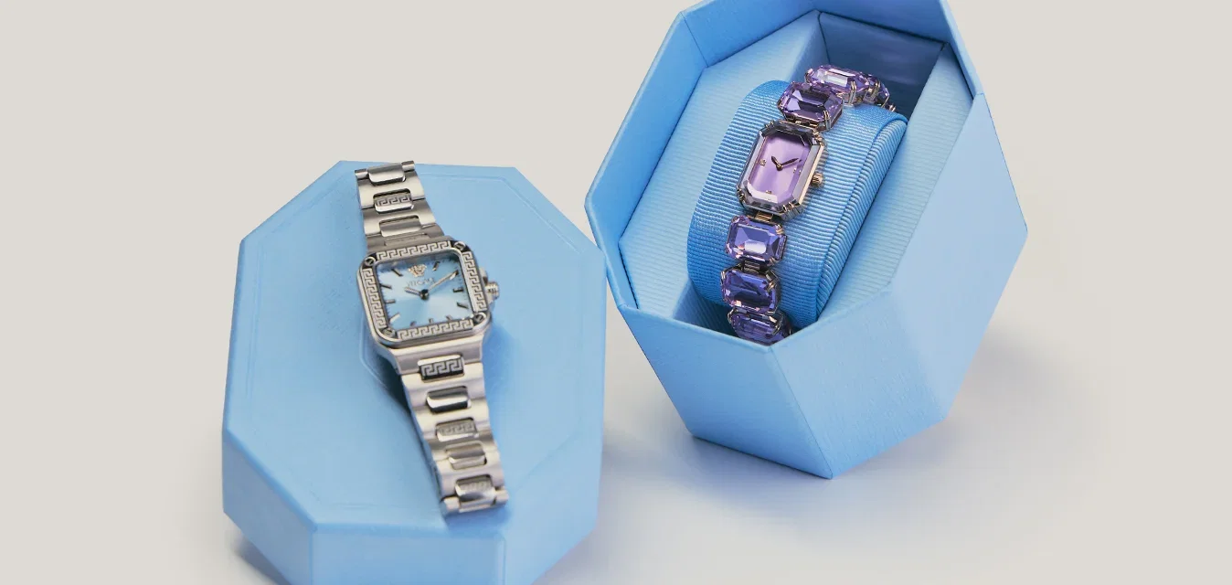 Due eleganti orologi da polso, uno argento e uno viola, esposti su e all’interno di supporti di forma geometrica azzurri su uno sfondo semplice.