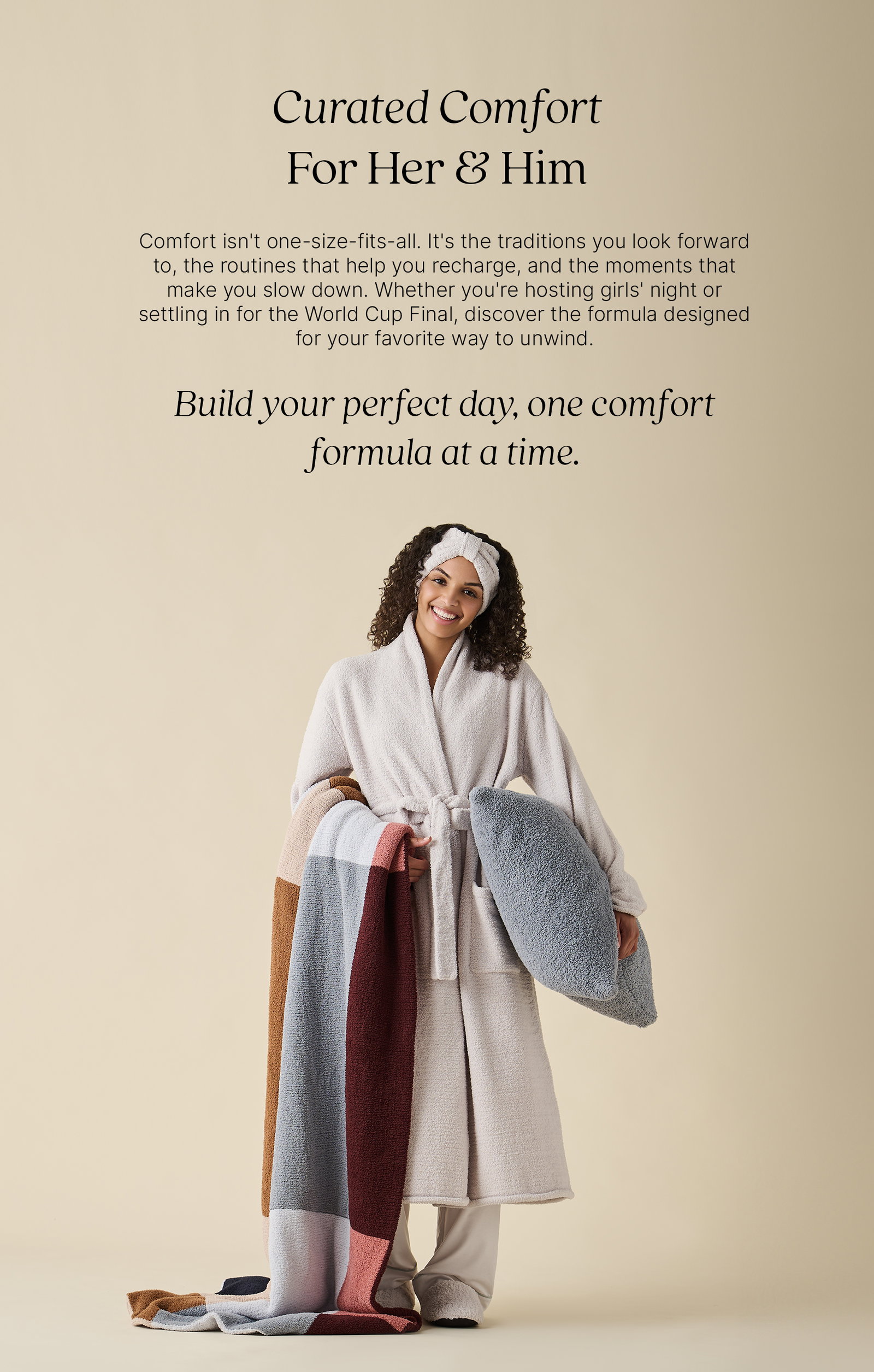 Curated Comfort For Her & Him. A model smiles while wearing a plush ivory Kashwére robe, matching headwrap, and cozy slippers. She holds a soft grey Cloud Pillow and a striped throw in warm neutral tones, standing against a soft cream background. Text reads: "Comfort isn't one-size-fits-all. It's the traditions you look forward to, the routines that help you recharge, and the moments that make you slow down. Whether you're hosting girls' night or settling in for the World Cup Final, discover the formula designed for your favorite way to unwind." Below, the headline reads: "Build your perfect day, one comfort formula at a time."