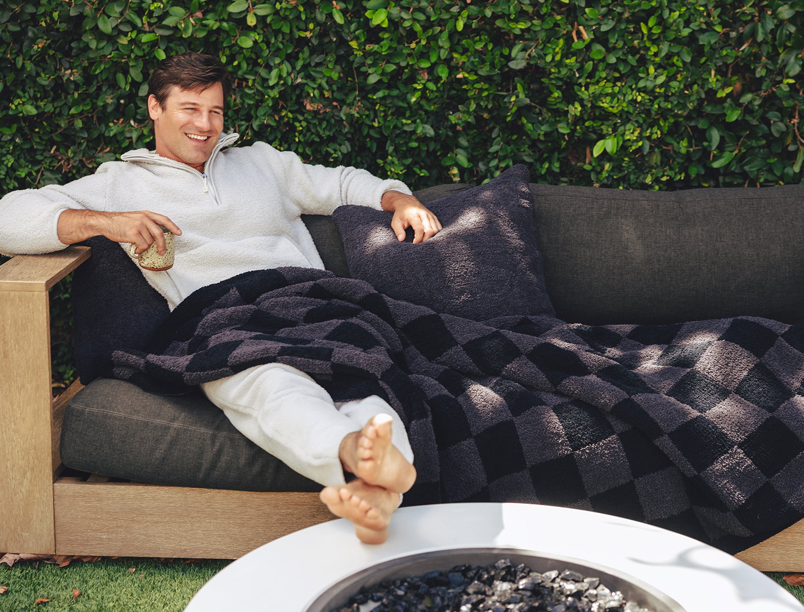A man relaxes on an outdoor sofa wearing a white Kashwére unisex half-zip pullover and matching joggers. He is wrapped in a black and charcoal check throw with coordinating plush pillows while holding a drink beside a modern fire pit. The cozy backyard setting highlights the warmth and comfort of the collection.