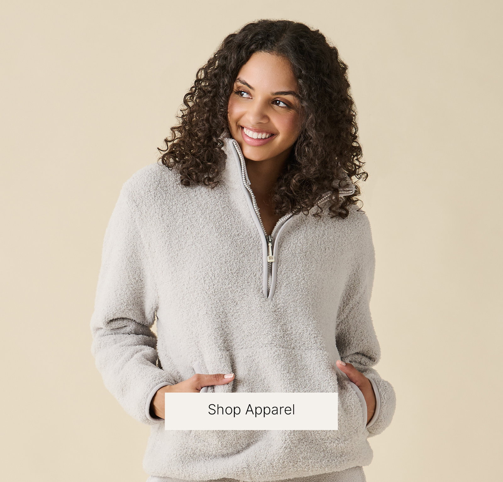 A smiling model wears the Kashwére unisex half-zip pullover in a soft neutral color, featuring a stand collar, quarter-zip neckline, and front kangaroo pocket. She poses against a warm cream background with the text, "Shop Apparel."