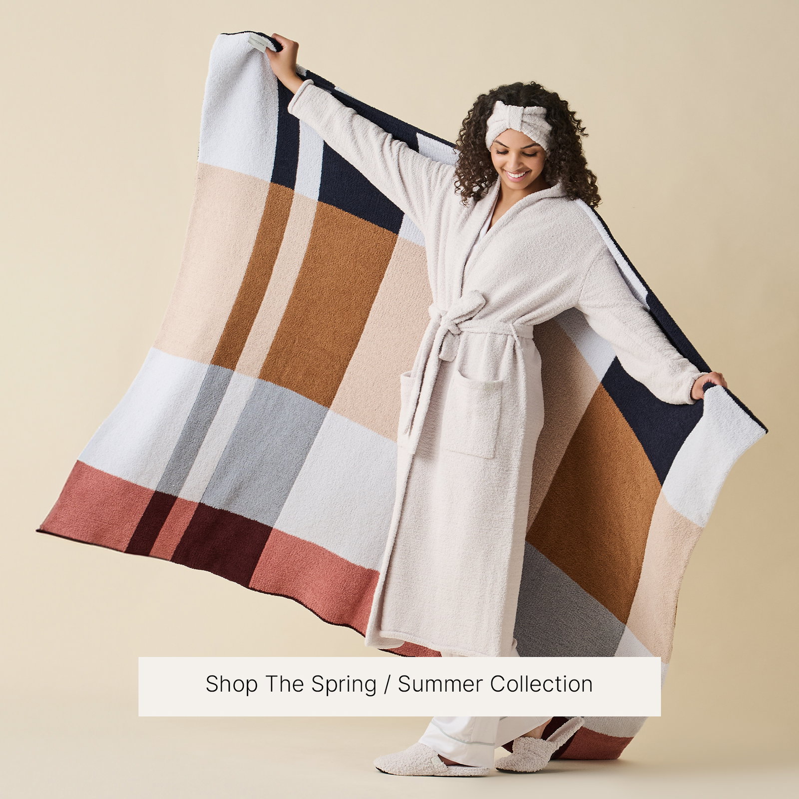 A smiling model wears an ivory Kashwére robe, matching plush headwrap, and cozy slippers while holding the reversible Plaid 2.0 Throw open behind her like a cape. The throw features oversized plaid in Dark Navy, Maliblu, Chestnut, Wheat, Ice Blue, Rosewood, and Syrah. Text at the bottom reads, "Shop The Spring / Summer Collection."