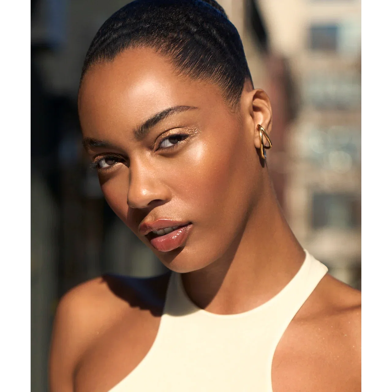 Close-up portrait of a woman with glowing skin and dark, slicked-back hair, against a blurred urban background.