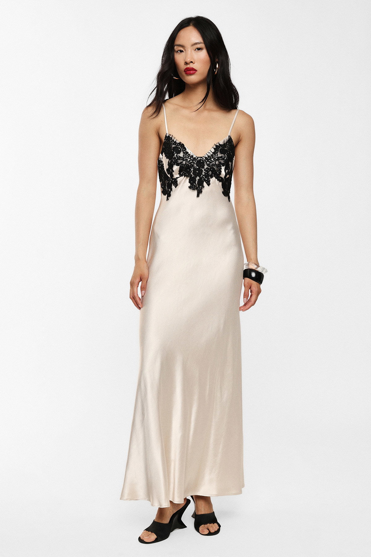 Woman modelling cream satin slip dress with black lace bodice and thin straps, wearing black sandals and bracelets on a plain studio background
