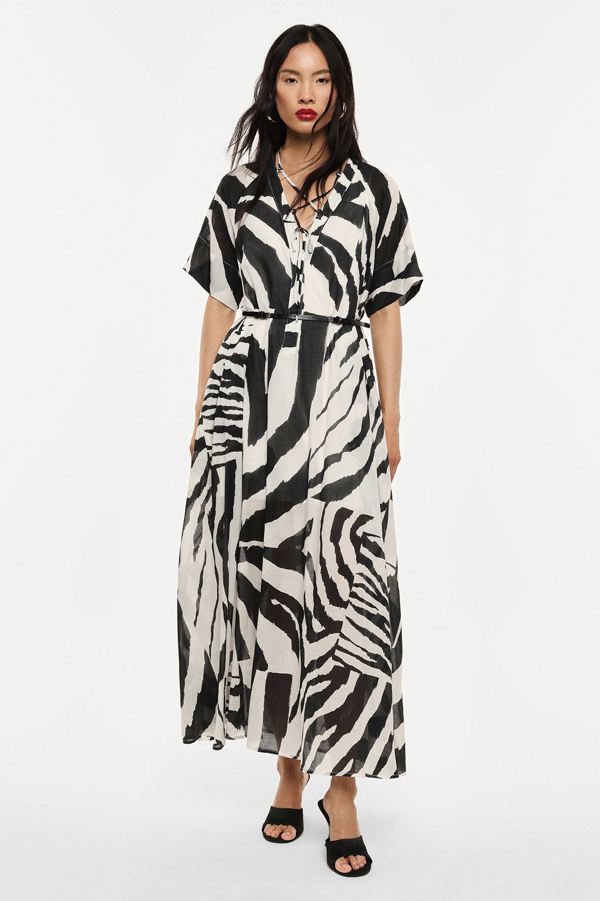 Woman wearing black-and-white zebra-print maxi dress with tie waist and short sleeves, standing in sandals against white background