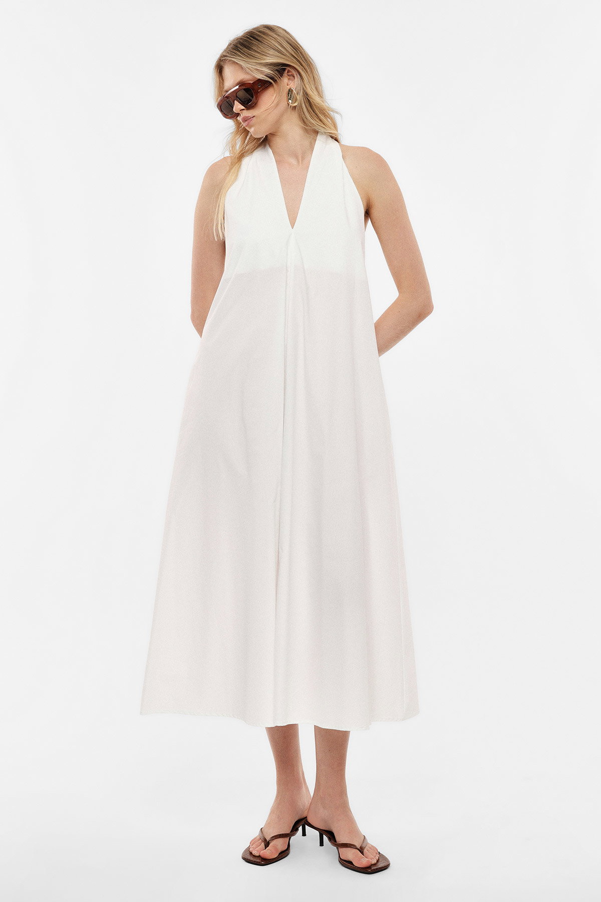 Woman in white halter midi dress with V-neck, sunglasses and heeled sandals, hands in pockets on white background