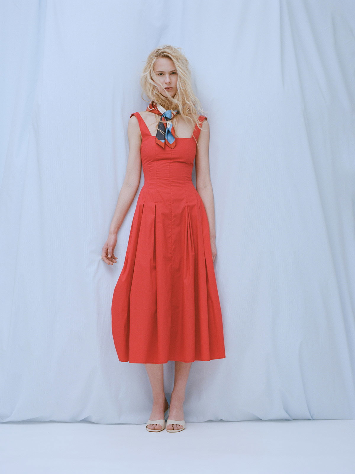 Blonde woman wearing a fitted red midi dress and patterned neck scarf standing against a pale draped backdrop
