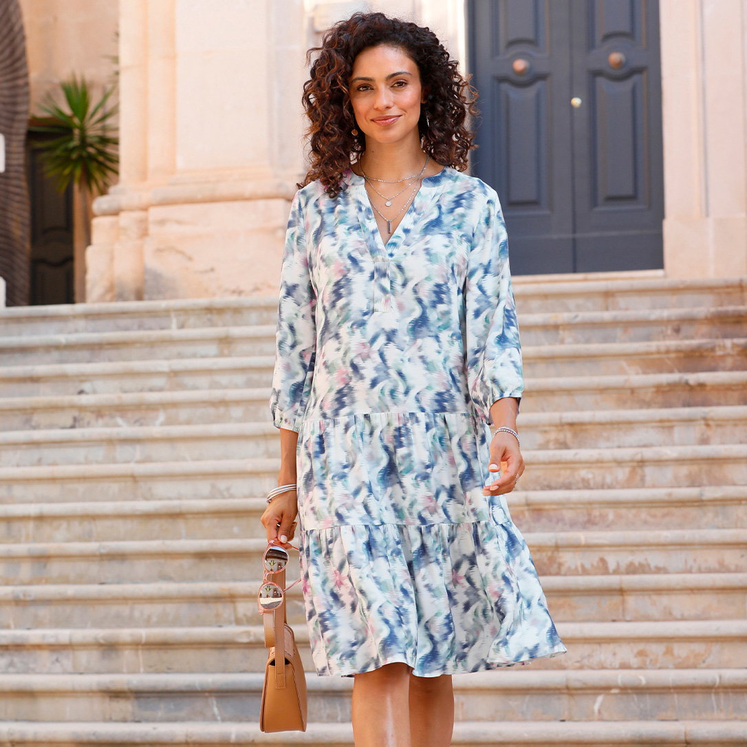 Ein Model mit lockigem Haar geht eine Treppe hinunter, trägt ein blau-weiß gemustertes Kleid mit 3/4-Ärmeln und hält eine braune Tasche.