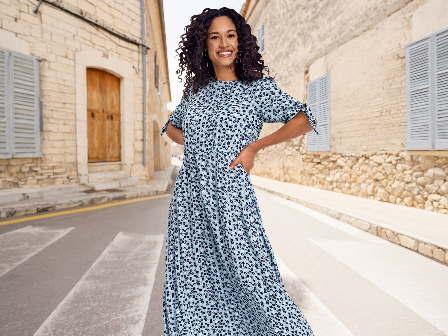 Ein Model mit lockigem Haar steht auf einer gepflasterten Straße, trägt ein blaues, gemustertes Kleid mit kurzen Ärmeln und lächelt.