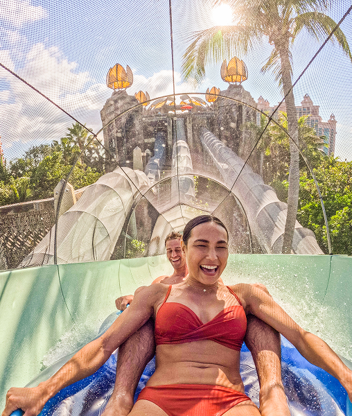 A laughing man and woman ride an inner tube together through an enclosed water slide tunnel.