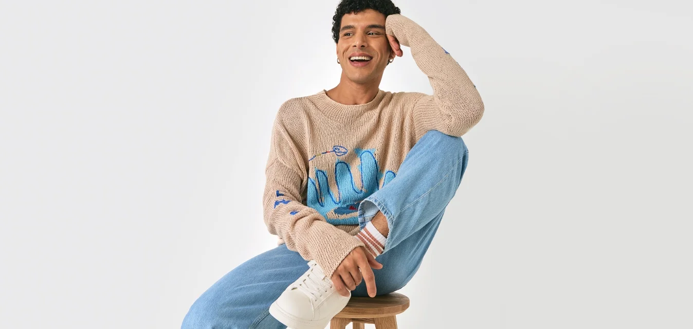 Jonge man die glimlacht, zittend met gekruiste benen op een houten kruk, gekleed in een beige trui met een blauw ontwerp en lichtblauwe spijkerbroek.