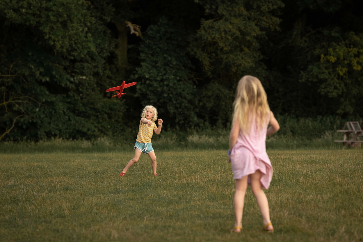 Two children play with a red toy airplane in a grassy field.