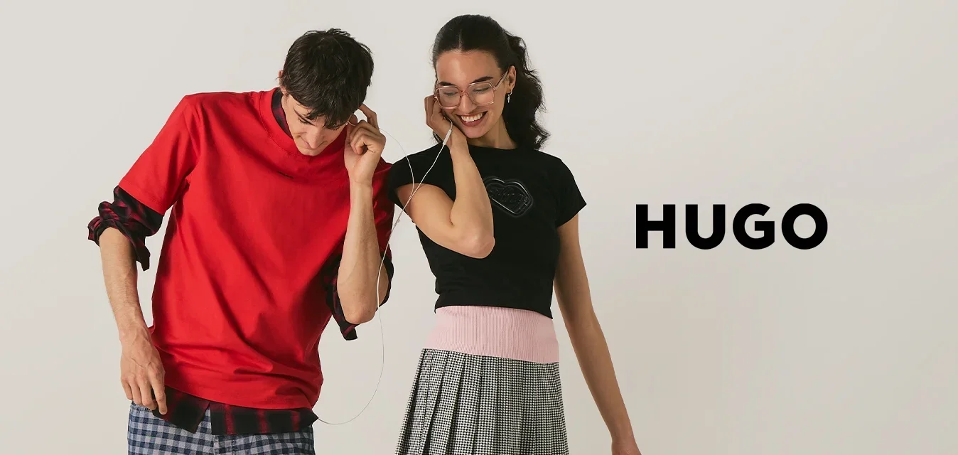 Adolescent garçon et fille partageant des écouteurs, debout proches, souriants ; il porte une chemise rouge et un pantalon à carreaux, elle porte un haut noir et une jupe grise.