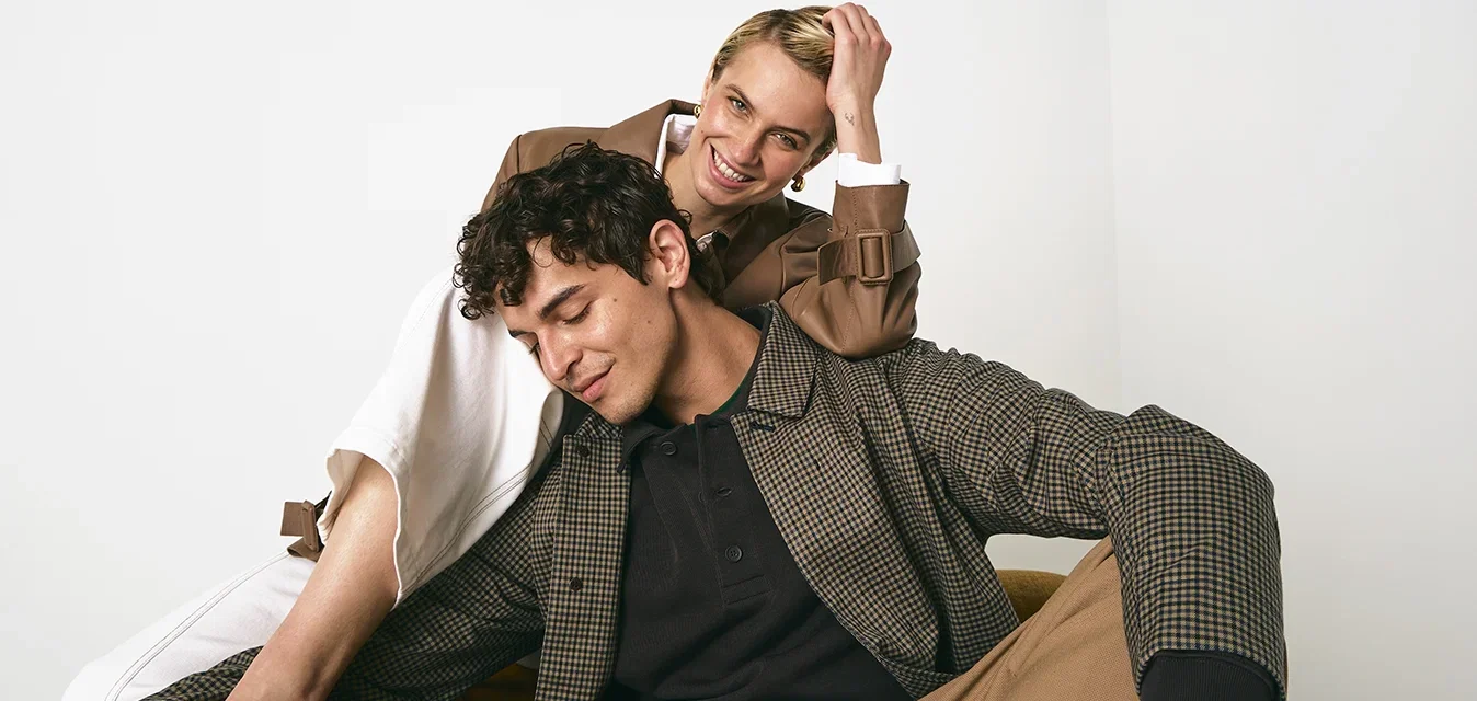 Deux modèles sont assis dans une chaise brune. L'un porte une chemise à carreaux, un polo noir et un pantalon beige ; l'autre est en manteau brun sur une robe blanche.