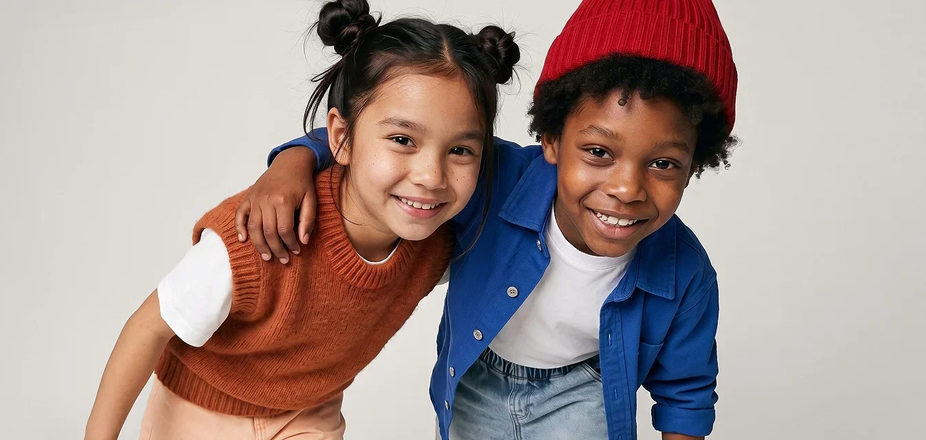 Dos niños sonrientes, uno con un gorro rojo y chaqueta azul, el otro con un chaleco de punto color óxido, inclinados hacia adelante con los brazos alrededor del otro.
