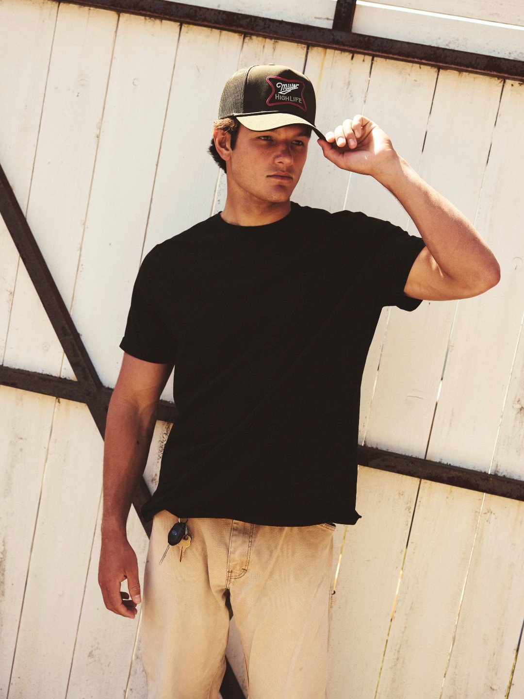 A man wearing a black t-shirt and tan pants standing outdoors against a white slatted fence, adjusting the brim of a dark green Miller High Life Fremont hat.