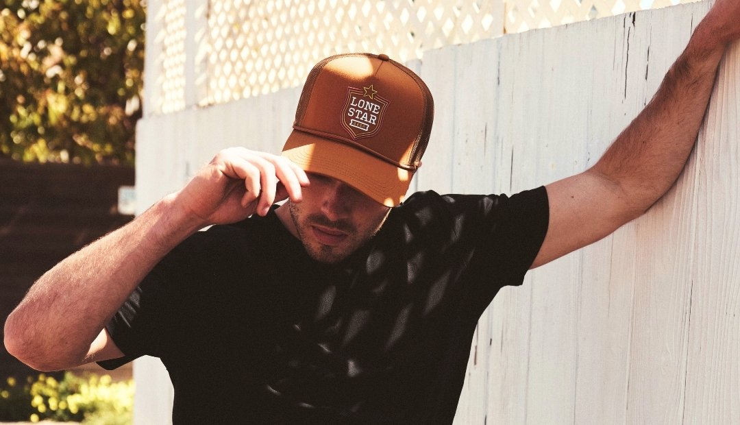 A man wearing a black t-shirt leans his arm against a white lattice fence, looking down while adjusting the brim of a brown Lone Star Fremont hat.