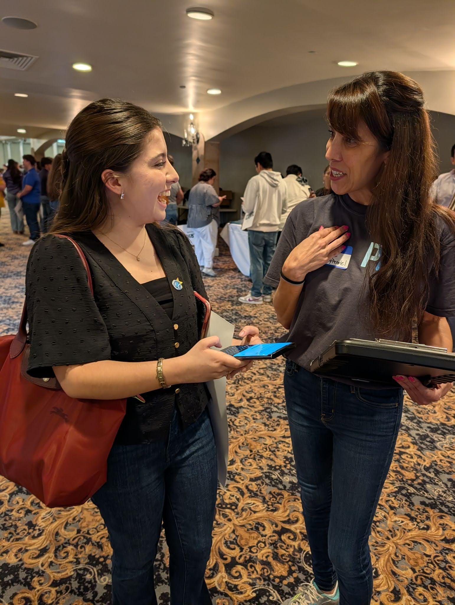 A woman wearing a black cardigan carrying a red purse holding her phone (left) laughing with a PxP volunteer holding a clipboard (right)