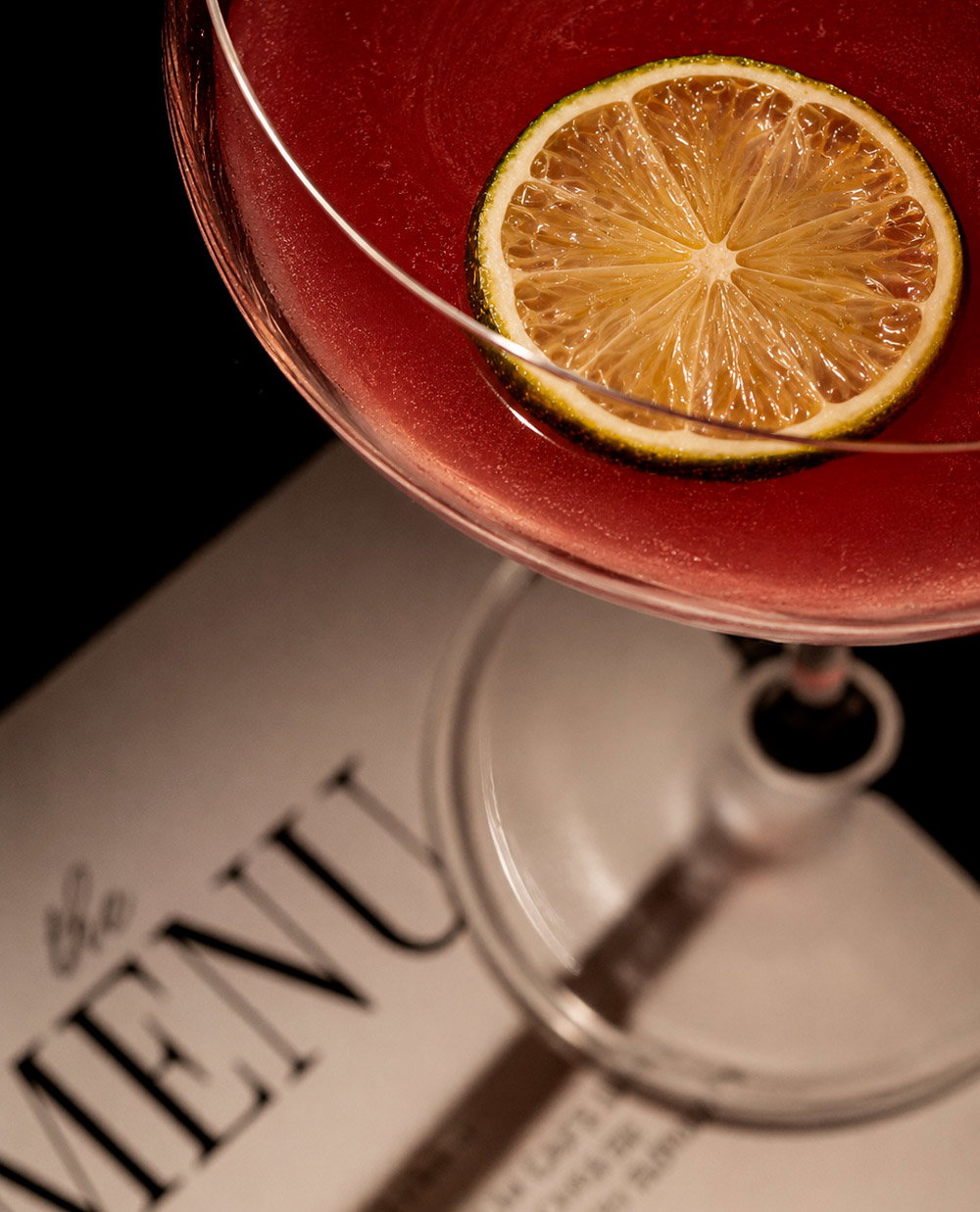 Top-down close-up of a red cocktail in a stemmed glass garnished with a lime wheel resting on a printed menu