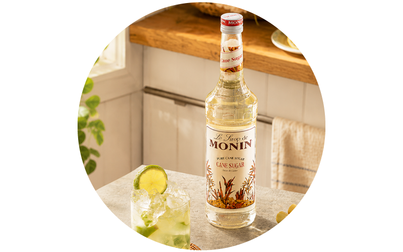 Bottle of Monin cane sugar syrup on a kitchen counter beside a lime-and-ice cocktail, natural light and wooden shelf background