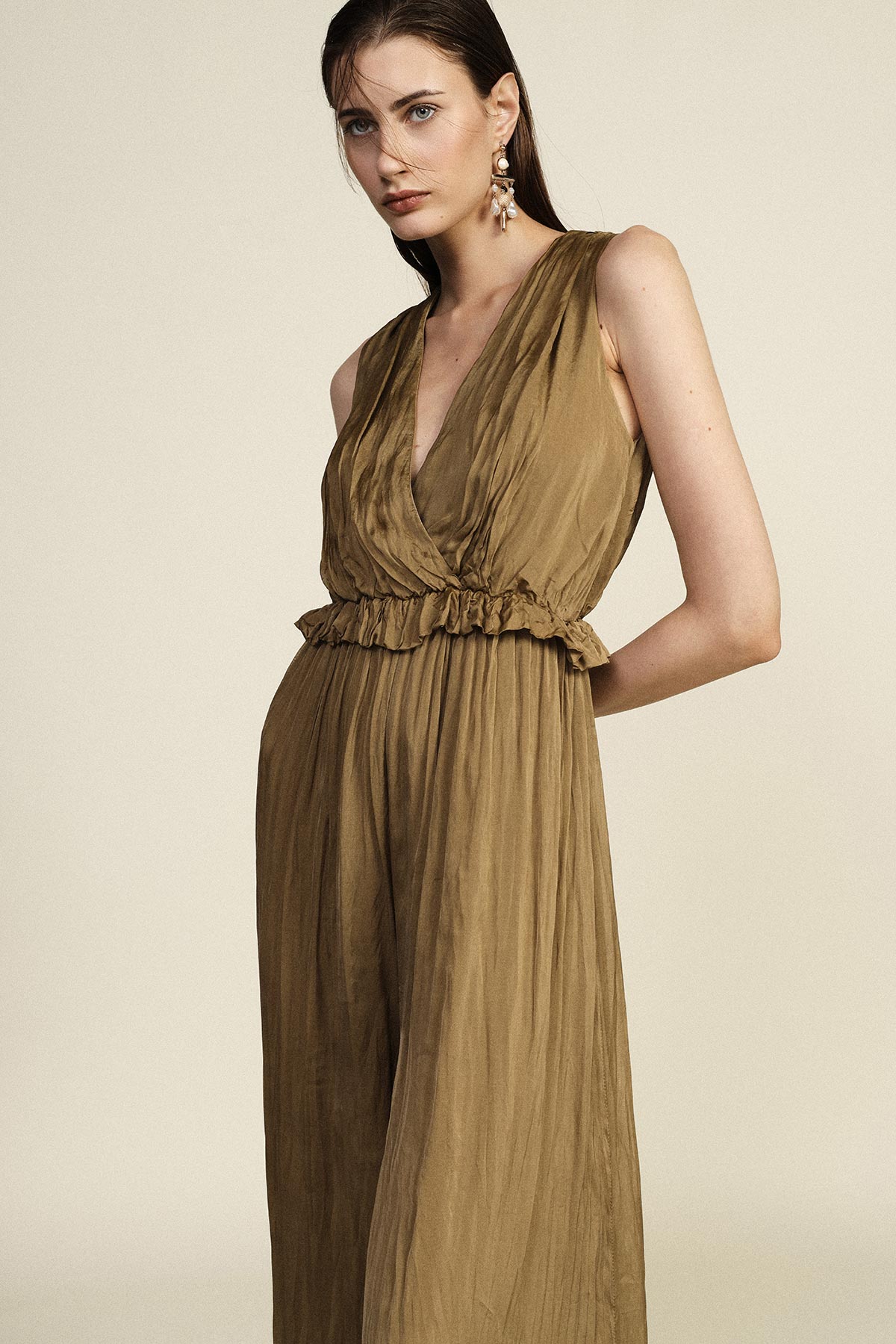 Woman modelling sleeveless olive-brown jumpsuit with deep V-neck, ruffled waist and wide legs, wearing statement earrings