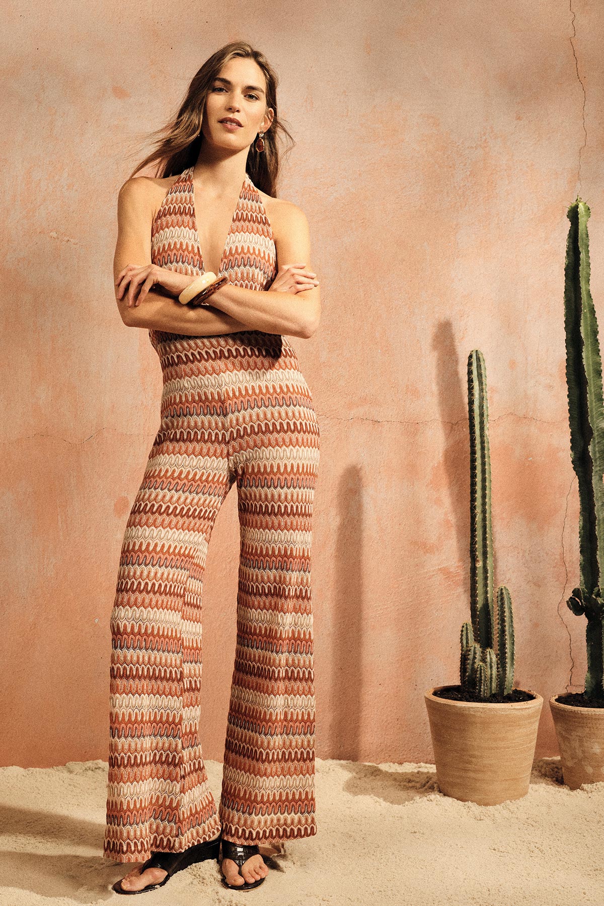 Woman in patterned halter-neck wide-leg jumpsuit and sandals, standing by potted cacti against a pink textured wall