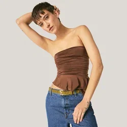 Vrouw met een bruin strapless topje en blauwe jeans leunt naar één kant, waarbij ze één arm op haar hoofd laat rusten tegen een effen achtergrond.